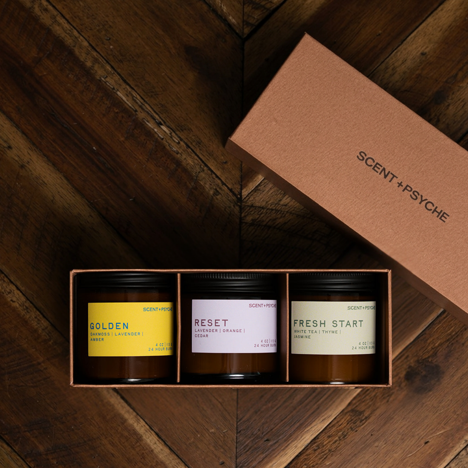 Scent + Psyche Energizing + Fresh soy candle discovery bundle in custom gift box. A three-scent candle gift set by Scent + Psyche in a brown elevated gift  box, displayed on a dark wood background with the matching lid leaning against it. The candles have black lids and labels in yellow, lavender, and sage green.