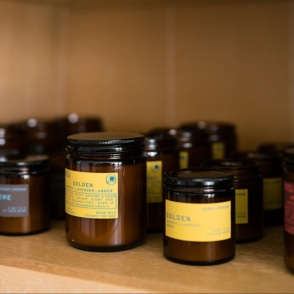 Amber glass candle jars with black lids and yellow labels arranged on wooden shelves, with one jar in sharp focus in the foreground.
