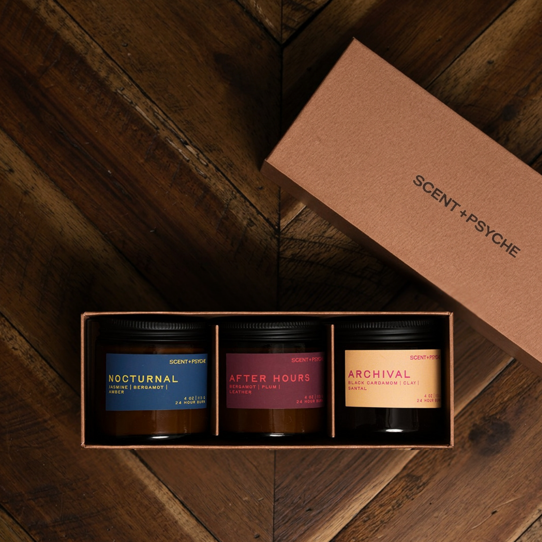 Warm + Floral Scented Candle Bundle | Discovery Size. A three-scented candle gift set by Scent+Psyche in a brown cardboard tray box, displayed on a dark wood chevron floor with the matching lid resting beside it. The candles feature dark amber glass jars with black lids and labels in navy blue, dusty rose, and warm tan.