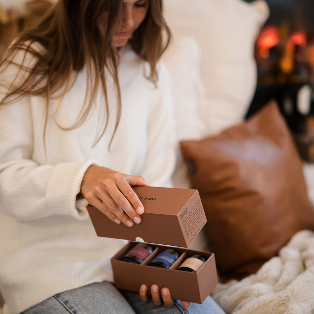 Warm + Floral Scented Candle Bundle | Discovery Size. A woman in a cozy living room setting, sitting on a couch and holding open a brown gift box containing several colorful packaged candles