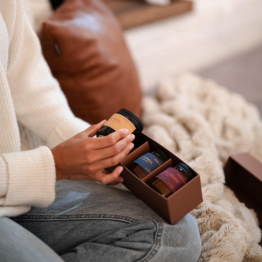 Warm + Floral Scented Candle Bundle | Discovery Size. A person sitting in a cozy living room by a fireplace, holding a small brown gift box containing several candles