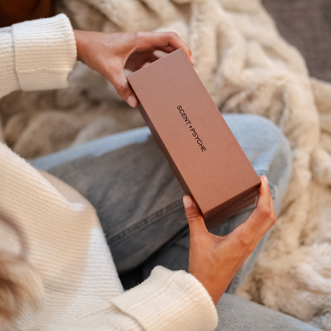A person in a cream knit sweater holds a brown rectangular box with the brand name Scent + Psyche  