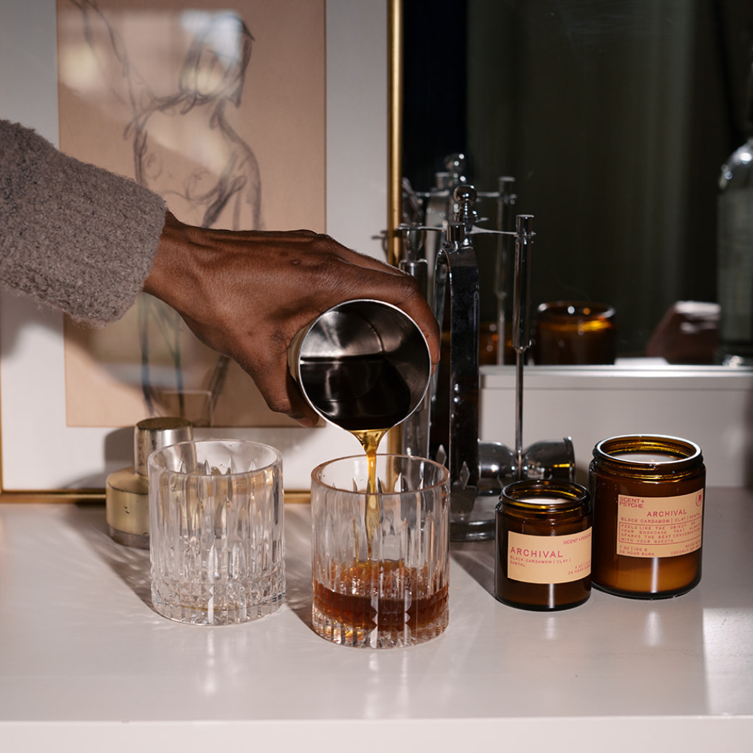 A hand pouring amber liquid from a metal pitcher into a faceted clear glass on a white tabletop, with amber and clear glass jars and a mirror in the background.