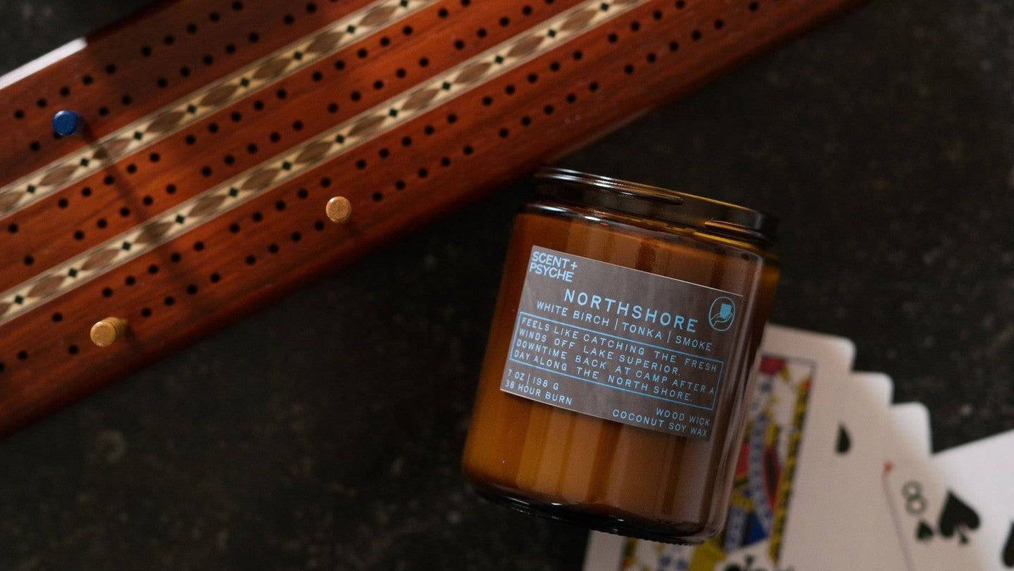 A styled flat-lay featuring a Northshore scented candle in an amber glass jar next to a wooden cribbage board, with playing cards fanned out on a dark textured surface.